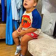 Andréa participe au concours pour gagner de l'argent avec cette photo : child, boy, blonde_hair, sitting, indoor, tshirt, shorts, sneakers, bench, clothing_rack, floor, looking_up, casual_clothing, young, curious, portrait, footwear, sock, wooden_floor, textile
