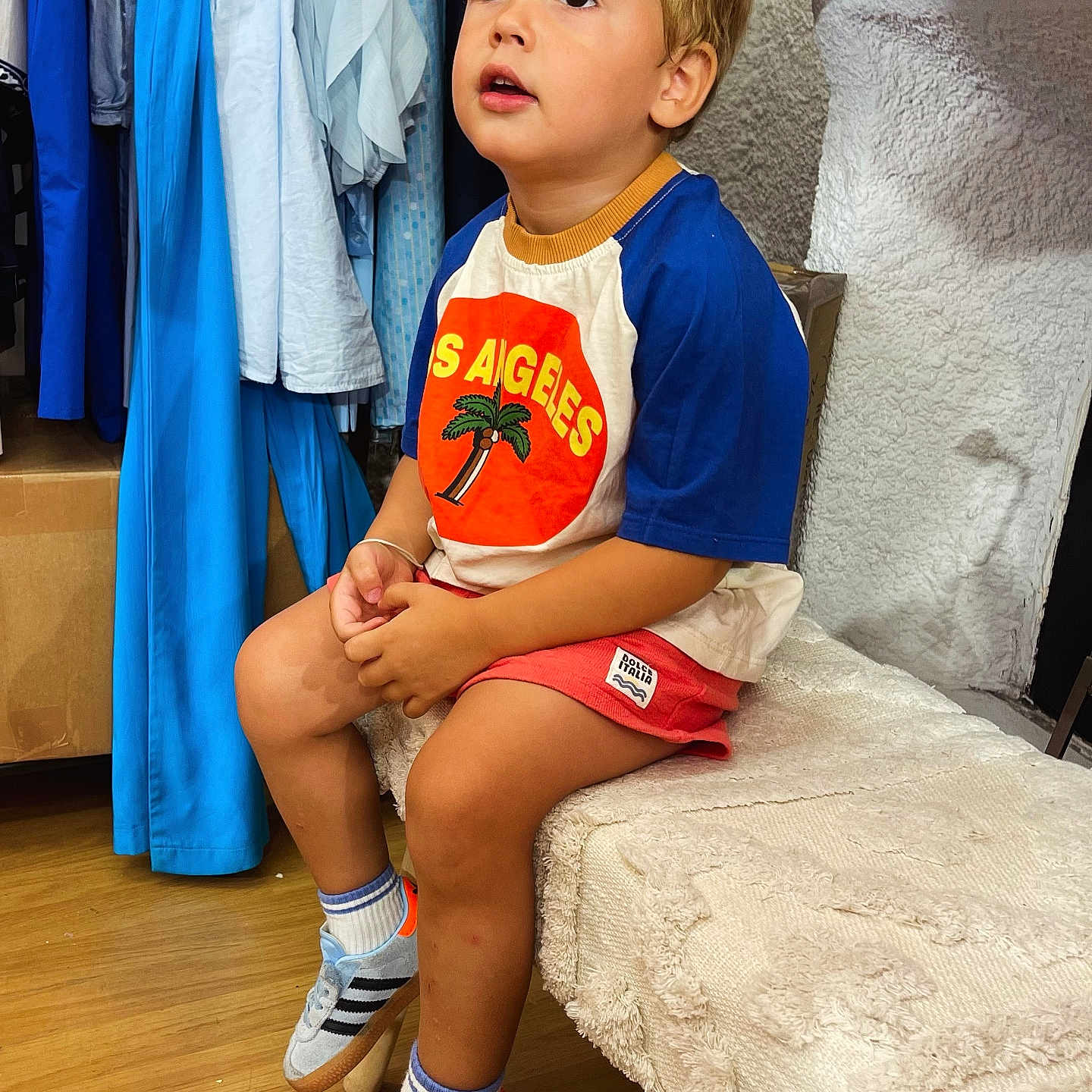 Andréa participe au concours pour gagner de l'argent avec cette photo : bench, blonde_hair, boy, casual_clothing, child, clothing_rack, curious, floor, footwear, indoor, looking_up, portrait, shorts, sitting, sneakers, sock, textile, tshirt, wooden_floor, young