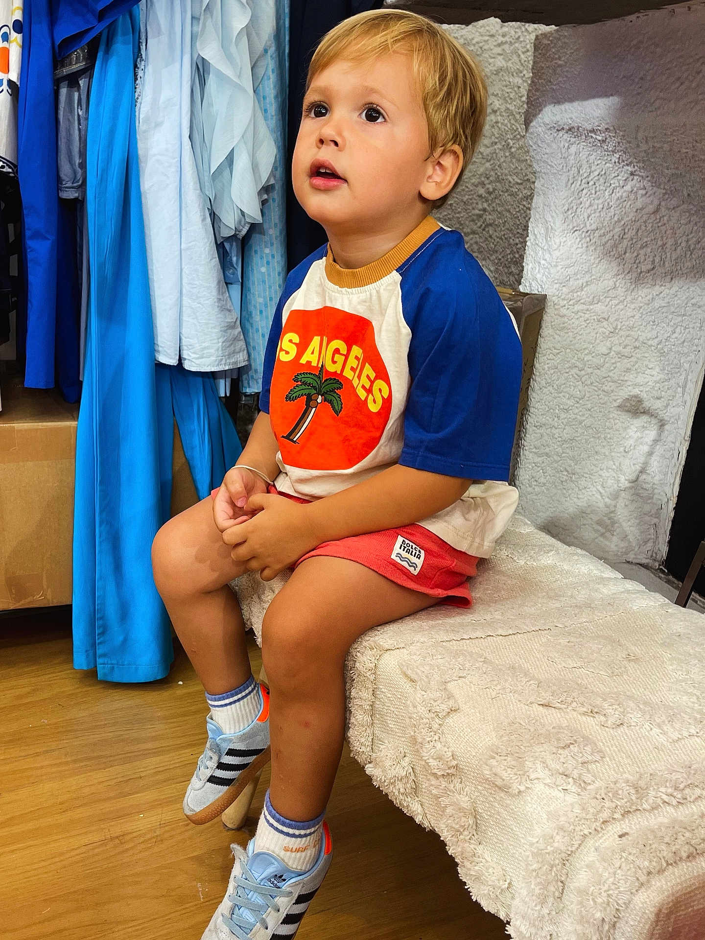 Andréa participe au concours pour gagner de l'argent avec cette photo : child, boy, blonde_hair, sitting, indoor, tshirt, shorts, sneakers, bench, clothing_rack, floor, looking_up, casual_clothing, young, curious, portrait, footwear, sock, wooden_floor, textile