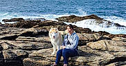 Inuk a rejoint le concours — aidez-le/la à gagner de superbes lots ! person, dog, rock, ocean, water, sky, cloud, outdoor, nature, coast, wave, animal, pet, casual_clothing, sitting, landscape, daytime, leisure, scenic, relaxation