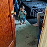 toddler, child, barefoot, indoors, cord, vacuum_cleaner, bed, person, poster, door, floor, carpet, furniture, sitting, clothing, handheld_object, room, technology, adult, casual