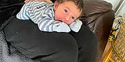 Xavier-James is registered to the contest to win money with this photo: baby, infant, person, clothing, striped_onesie, lap, indoor, couch, leather, basket, floor, wooden_floor, hand, face, curious, resting, black_clothing, blanket, home, casual