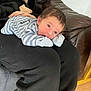 baby, infant, person, clothing, striped_onesie, lap, indoor, couch, leather, basket, floor, wooden_floor, hand, face, curious, resting, black_clothing, blanket, home, casual