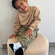 Eyden participe au concours pour gagner de l'argent avec cette photo : beige_sweatshirt, candid, casual_clothing, child, curly_hair, floor_tiles, happy, indoor, joyful, light_wall, olive_pants, person, playful, portrait, pouf, seated, smiling, tilted_head, white_sneakers, young
