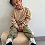 Eyden participe au concours pour gagner de l'argent avec cette photo : beige_sweater, casual_clothing, child, curly_hair, cute, floor, green_pants, happy, indoor, light, ottoman, person, playful, portrait, relaxed, sitting, smiling, wall, white_sneakers, young_child