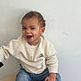 bracelet, casual_clothing, child, curly_hair, cute, face, happy, indoor, jeans, joyful, light_background, person, portrait, real_photo, sitting, smiling, sweater, toddler, white_wall, young_child
