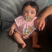 Selina joined the competition — help win amazing prizes! baby, pacifier, pink_clothing, sitting, gray_cushion, chair, adult_hand, indoor, cute, infant, portrait, child, people, comfort, relaxed, colorful, floral_pattern, bird_design, soft_lighting, closeup