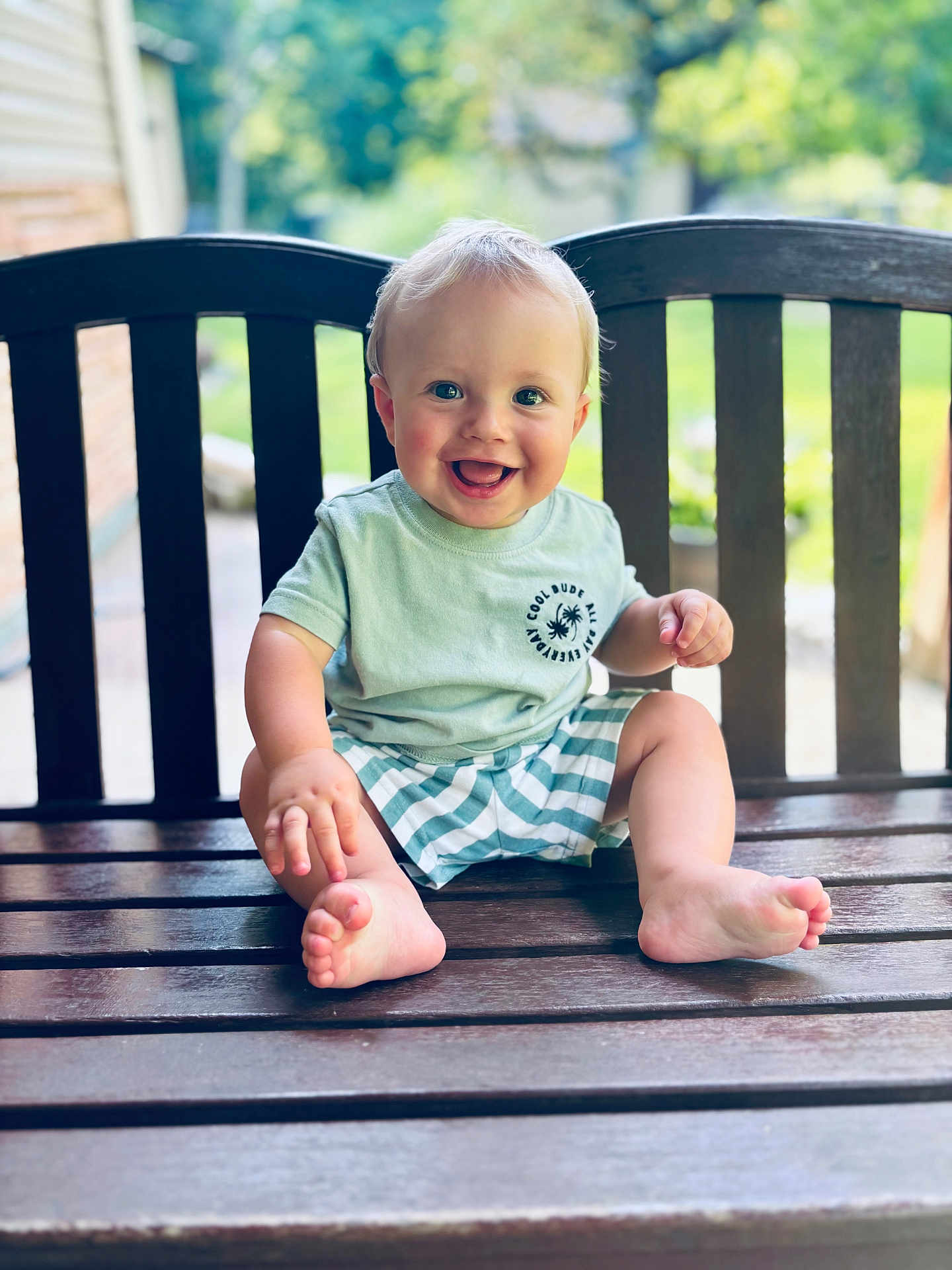 Finnley is registered to the contest to win money with this photo: baby, child, smiling, barefoot, outdoor, bench, wooden_bench, green_shirt, striped_shorts, happy, portrait, cute, sitting, young_child, daylight, nature, grass, blurred_background, person, summer