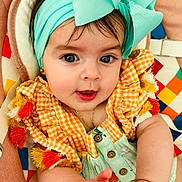 Esmeralda is registered to the contest to win money with this photo: baby, child, infant, portrait, smiling, indoors, headband, bow, fashion, colorful, gingham, dress, tassels, cushion, blanket, lying_down, hands, eyes, jewelry, close_up