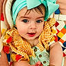 baby, child, infant, portrait, smiling, indoors, headband, bow, fashion, colorful, gingham, dress, tassels, cushion, blanket, lying_down, hands, eyes, jewelry, close_up
