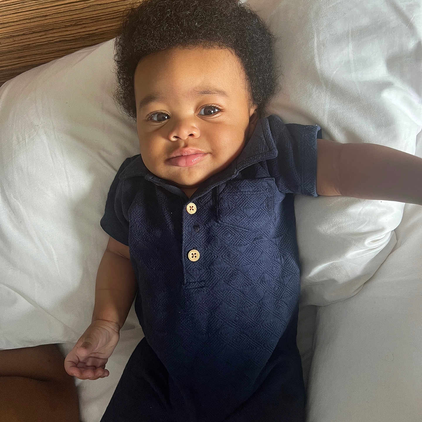 Ramarn is registered to the contest to win money with this photo: arm, baby, bed, child, comfort, curly_hair, cute, face, hand, indoors, infant, lying_down, navy_blue_clothing, portrait, relaxed, romper, skin, smile, white_pillows, wooden_buttons