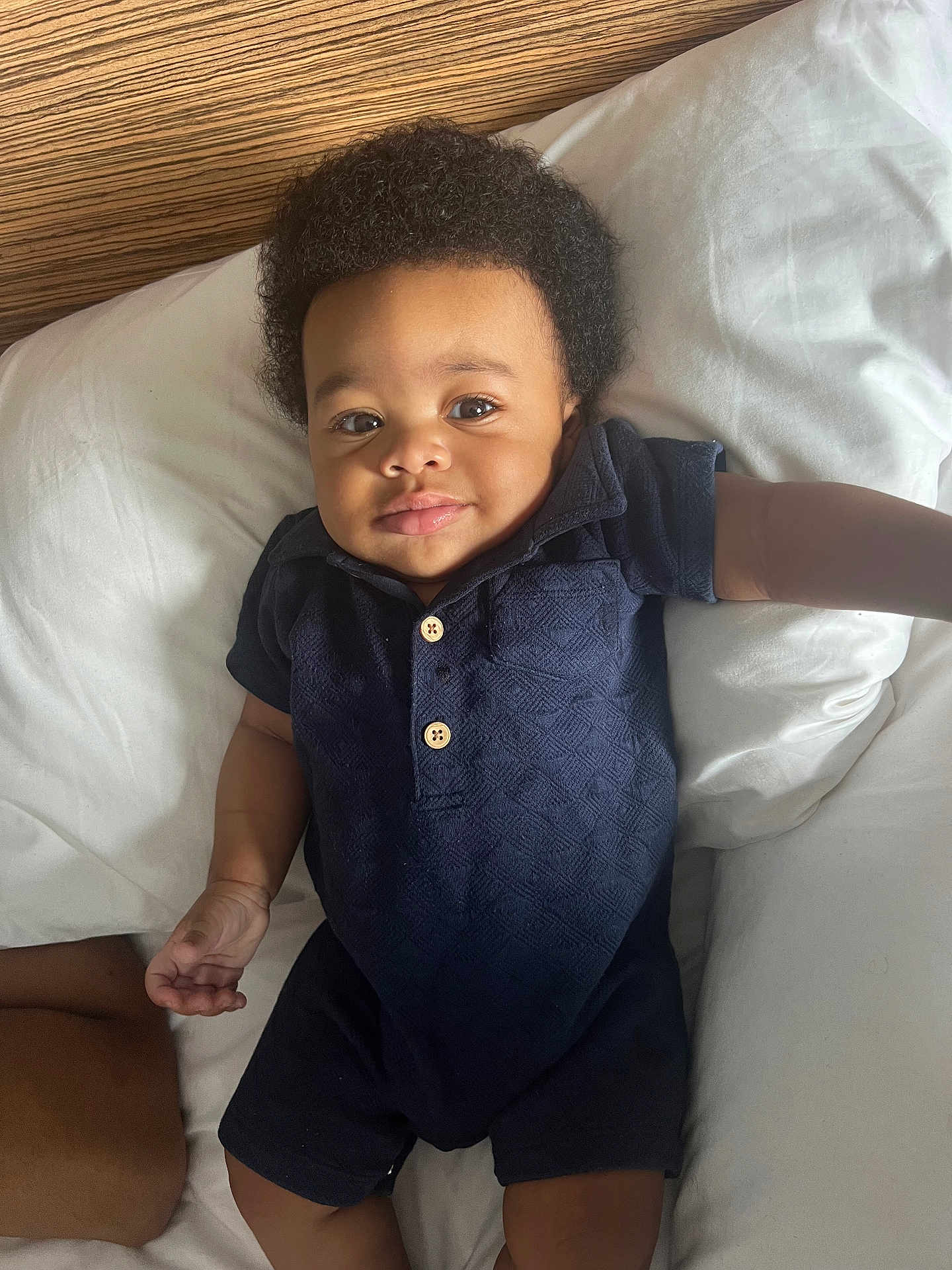 Ramarn is registered to the contest to win money with this photo: baby, infant, child, smile, curly_hair, navy_blue_clothing, romper, wooden_buttons, bed, white_pillows, relaxed, cute, portrait, indoors, skin, face, hand, arm, lying_down, comfort