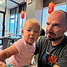baby, child, man, indoor, restaurant, lantern, window, table, chair, pink_shirt, facial_expression, holding, light, casual_clothing, person, portrait, curious, beard, short_hair, seated