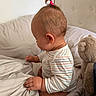 baby, child, toddler, bed, bedding, blanket, teddy_bear, stuffed_animal, striped_clothing, hair_tie, pony_tail, hand, ear, side_view, indoor, soft_light, curious, cozy, cushion, pajamas