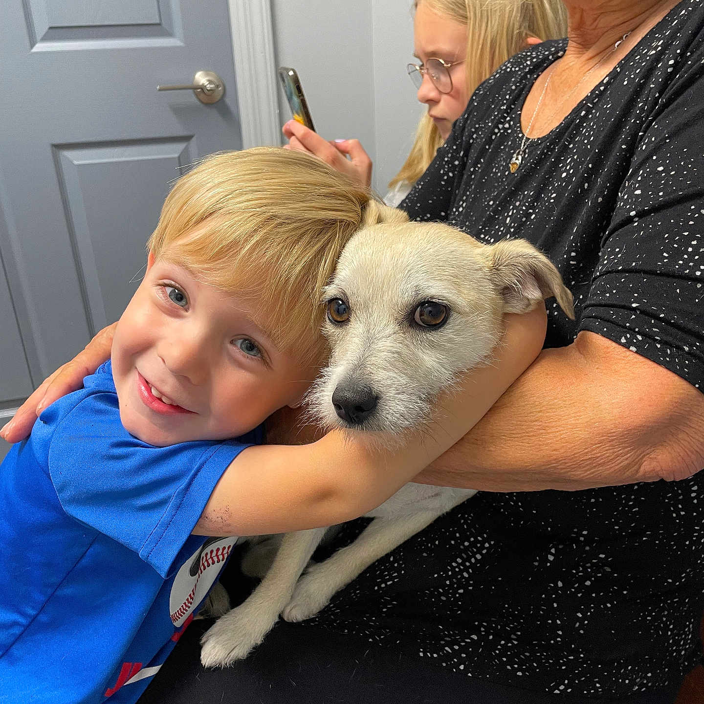 Sadie is registered to the contest to win money with this photo: affection, black_clothing, blond_hair, boy, candid, casual, child, closeup, dog, family, glasses, hug, indoor, pet, seated, smartphone, smile, warmth, white_dog, woman
