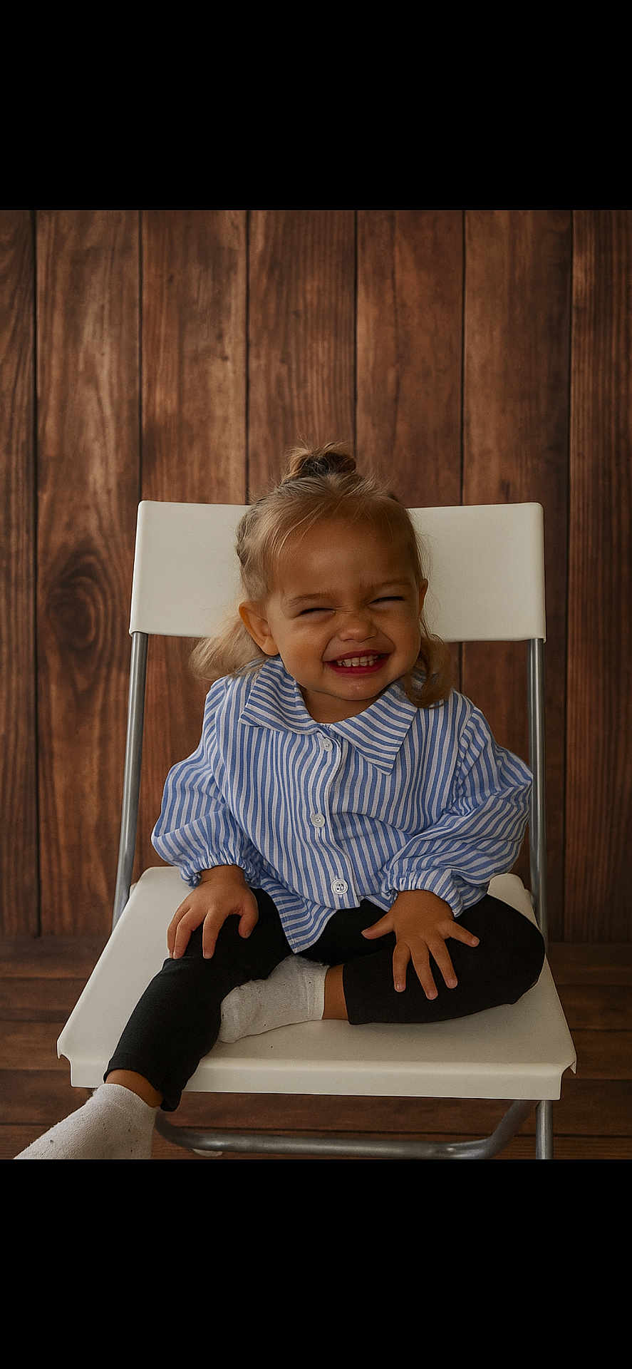 Malya participe au concours pour gagner de l'argent avec cette photo : child, toddler, smiling, chair, striped_shirt, black_pants, white_socks, wooden_background, indoor, portrait, cute, happy, sitting, cross_legged, young_child, casual_clothing, small_hands, blond_hair, playful, funny_face