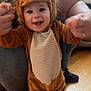 Mavis is registered to the contest to win money with this photo: baby, child, lion_costume, smiling, happy, person, indoor, floor, wooden_floor, couch, furniture, adult, holding_hands, costume, animal_outfit, footwear, cute, face, head, portrait