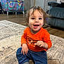 toddler, child, smiling, orange_shirt, blue_pants, sitting, rug, indoor, happy, person, floor, hair, eyes, long_sleeves, sock, home, carpet, furniture, door, playpen