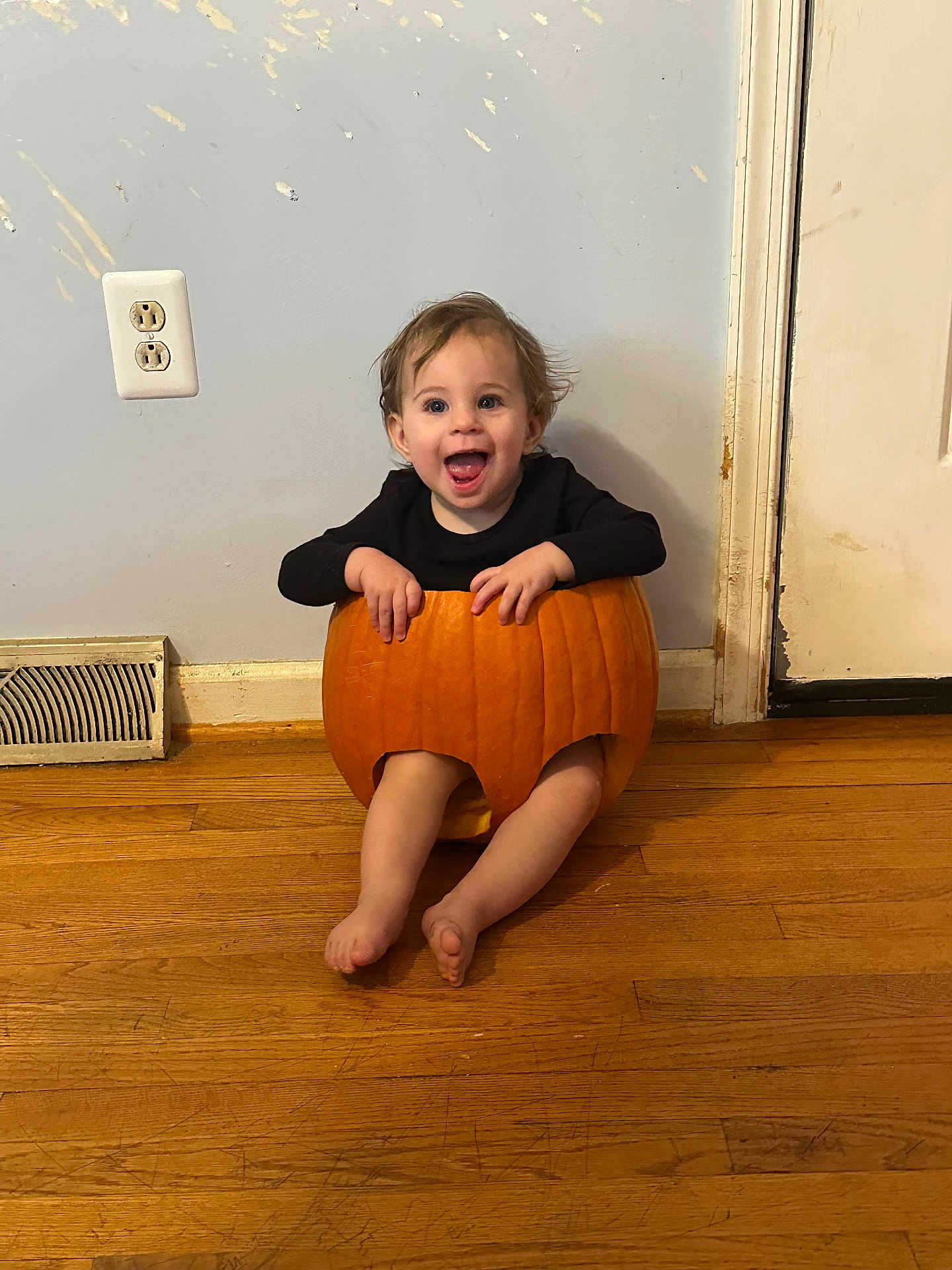 Mavis is registered to the contest to win money with this photo: baby, black_shirt, child, cute, door, face, feet, floor, hands, happy, holiday, indoor, orange, playful, pumpkin, sitting, smiling, toddler, wall, wooden_floor