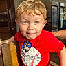 child, boy, red_shirt, baseball_graphic, blond_hair, blue_eyes, smile, indoor, chair, wood, kitchen, refrigerator, sunlight, shadow, person, casual_clothing, cute, portrait, playful, toddler