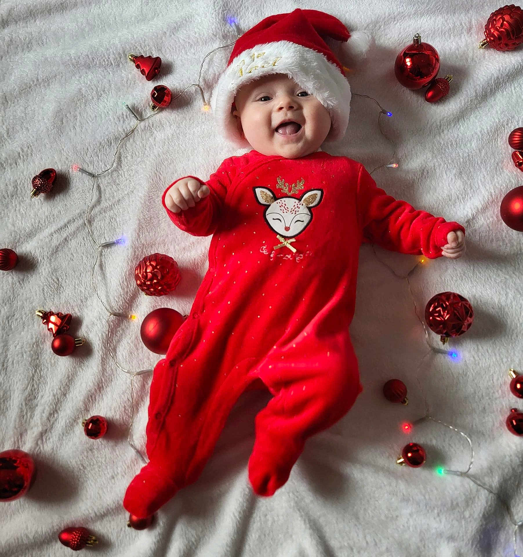 Nina a rejoint le concours — aidez-le/la à gagner de superbes lots ! baby, infant, smiling, red_clothing, christmas_hat, christmas_ornaments, holiday_lights, festive, cute, lying_down, white_blanket, celebration, joy, happy, seasonal, toddler, face, headwear, christmas, decoration