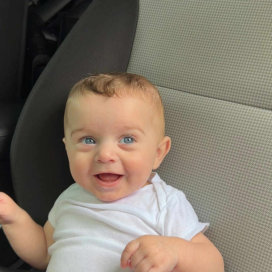 Micah joined the competition — help win amazing prizes! baby, child, car_seat, white_clothing, smiling, blue_eyes, happy, infant, cute, sitting, portrait, one_person, indoors, casual, human, young_child, cute_baby, car_interior, seat, clothing