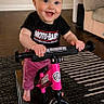 baby, child, toddler, smiling, balance_bike, strider, pink_bike, indoor, living_room, rug, hardwood_floor, sofa, hands, feet, blue_eyes, clothing, happy, play, toy, portrait