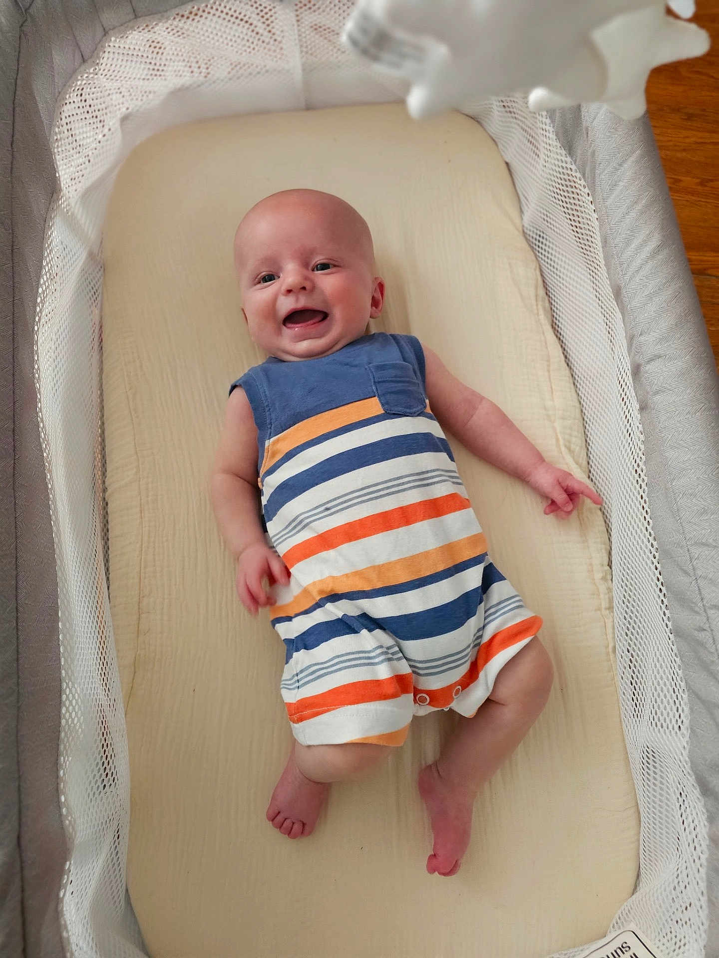Riley is registered to the contest to win money with this photo: baby, infant, smiling, romper, striped_clothing, bassinet, crib, bedding, mesh_sides, mobile, happy, child, indoors, flooring, wood_floor, cute, portrait, newborn, person, sleeping_area