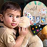 toddler, child, face, plush_toy, teddy_bear, party_hat, polka_dot, cuddling, portrait, indoors, soft_lighting, short_hair, brown_hair, skin, clothing, bracelet, happy, cute, celebration, background_blur