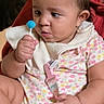 baby, child, lollipop, pacifier_clip, floral_onesie, bib, hand, face, curious, seated, red_background, skin, infant, expression, indoors, person, cute, young, holding, looking