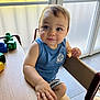 toddler, child, blue_eyes, snack, wooden_table, building_blocks, indoor, natural_light, curious_expression, blue_shirt, striped_shorts, high_chair, frosted_window, hand, face, person, furniture, play, casual_clothing, daylight