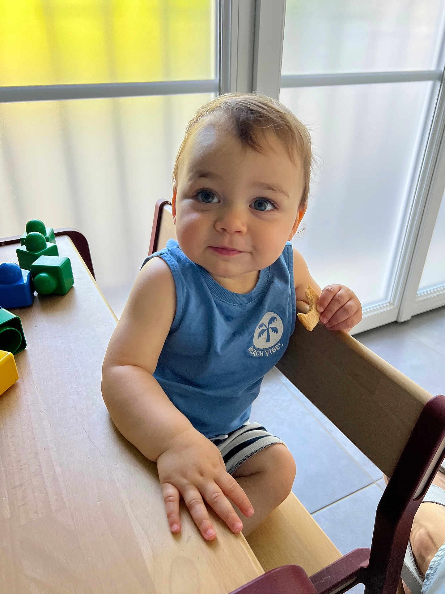 énoha a rejoint le concours — aidez-le/la à gagner de superbes lots ! toddler, child, blue_eyes, snack, wooden_table, building_blocks, indoor, natural_light, curious_expression, blue_shirt, striped_shorts, high_chair, frosted_window, hand, face, person, furniture, play, casual_clothing, daylight