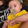 Jerry is registered to the contest to win money with this photo: baby, pacifier, person, yellow_shirt, indoor, curious_expression, holding, arm, face, closeup, child, casual_clothing, text_on_shirt, toddler, human, skin, hair, seated, couch, comfort