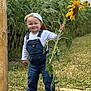 Carson is registered to the contest to win money with this photo: cap, casual_clothing, child, daylight, denim, field, flower, grass, greenery, happy, hat, nature, outdoor, overalls, plant, portrait, smile, sunflower, toddler, young_child