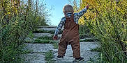 Carson is registered to the contest to win money with this photo: adventure, back_view, casual_clothing, child, daytime, exploring, grass, greenery, nature, outdoors, overalls, pathway, plaid_shirt, plants, sky, stone_path, sunlight, toddler, walking, young_child
