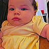 Zara is registered to the contest to win money with this photo: baby, child, infant, portrait, face, eyes, yellow_dress, yellow_clothing, sitting, indoor, home, furniture, adult_arm, close_up, looking_at_camera, serious_expression, curly_hair, colorful_background, photography, family