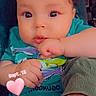baby, infant, child, face, eyes, wide_eyed, indoor, portrait, close_up, sitting, turquoise_shirt, clothing, hands, fist, adult_arm, heart_sticker, date_text, text_overlay, home, photograph
