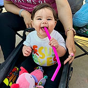 Isabella is registered to the contest to win money with this photo: baby, child, stroller, toy, pink_straw, smiling, happy, person, clothing, blanket, hand, outdoor, casual, supporting_hand, colorful, playful, cute, infant, sitting, daylight