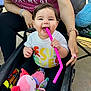 baby, child, stroller, toy, pink_straw, smiling, happy, person, clothing, blanket, hand, outdoor, casual, supporting_hand, colorful, playful, cute, infant, sitting, daylight