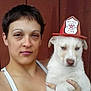 Odin is registered to the contest to win money with this photo: person, dog, white_dog, fire_chief_hat, pet, animal, portrait, serious_expression, short_hair, indoor, holding, looking_at_camera, brown_background, face, headwear, canine, human, companion, cute, close_up