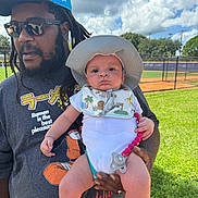 Vash joined the competition — help win amazing prizes! baby, infant, child, father, adult, portrait, outdoor, baseball_field, stadium, grass, sky, clouds, sun_hat, sunglasses, beard, dreadlocks, holding, pacifier, bib, smiling