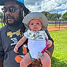 baby, infant, child, father, adult, portrait, outdoor, baseball_field, stadium, grass, sky, clouds, sun_hat, sunglasses, beard, dreadlocks, holding, pacifier, bib, smiling