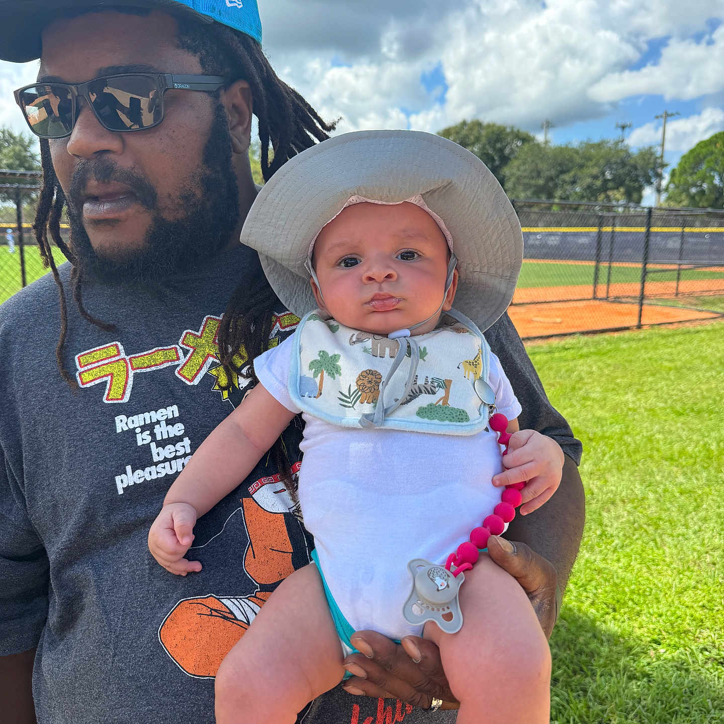 Vash joined the competition — help win amazing prizes! baby, infant, child, father, adult, portrait, outdoor, baseball_field, stadium, grass, sky, clouds, sun_hat, sunglasses, beard, dreadlocks, holding, pacifier, bib, smiling