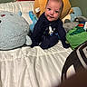 baby, plush_toys, bed, white_bedspread, blue_clothing, stuffed_shark, stuffed_bear, green_wall, child, smiling, sitting, cute, indoor, soft_toys, toddler, face, head, person, cozy, playful