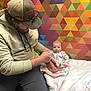 baby, infant, adult, man, bearded_man, cap, sunglasses, hoodie, onesie, blanket, colorful_wallpaper, geometric_pattern, triangle_pattern, chair, sitting, indoor, portrait, holding, family, bonding