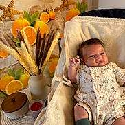 Mani participe au concours pour gagner de l'argent avec cette photo : baby, infant, blanket, romper, green_socks, hand, face, table, vase, dried_grasses, candle, container, orange_slices, starfish, decor, indoor, cozy, expression, patterned_clothing, soft_texture
