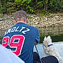animal, back_view, blue_shirt, boat, canine, collar, dog, fishing, leisure, man, nature, outdoor, pet, recreation, riverbank, rocks, scenery, summer, trees, water