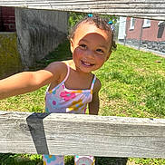 Kiya is registered to the contest to win money with this photo: child, toddler, smile, outdoor, grass, wooden_fence, sunlight, colorful_clothing, hair_ties, happy, playful, shoes, person, daylight, greenery, flower_pattern, cute, portrait, summer, casual