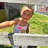 child, toddler, smile, outdoor, grass, wooden_fence, sunlight, colorful_clothing, hair_ties, happy, playful, shoes, person, daylight, greenery, flower_pattern, cute, portrait, summer, casual