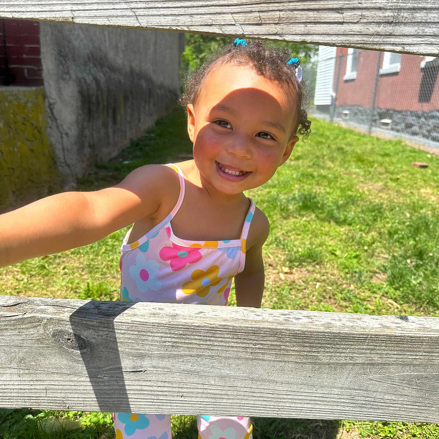 Kiya is registered to the contest to win money with this photo: child, toddler, smile, outdoor, grass, wooden_fence, sunlight, colorful_clothing, hair_ties, happy, playful, shoes, person, daylight, greenery, flower_pattern, cute, portrait, summer, casual
